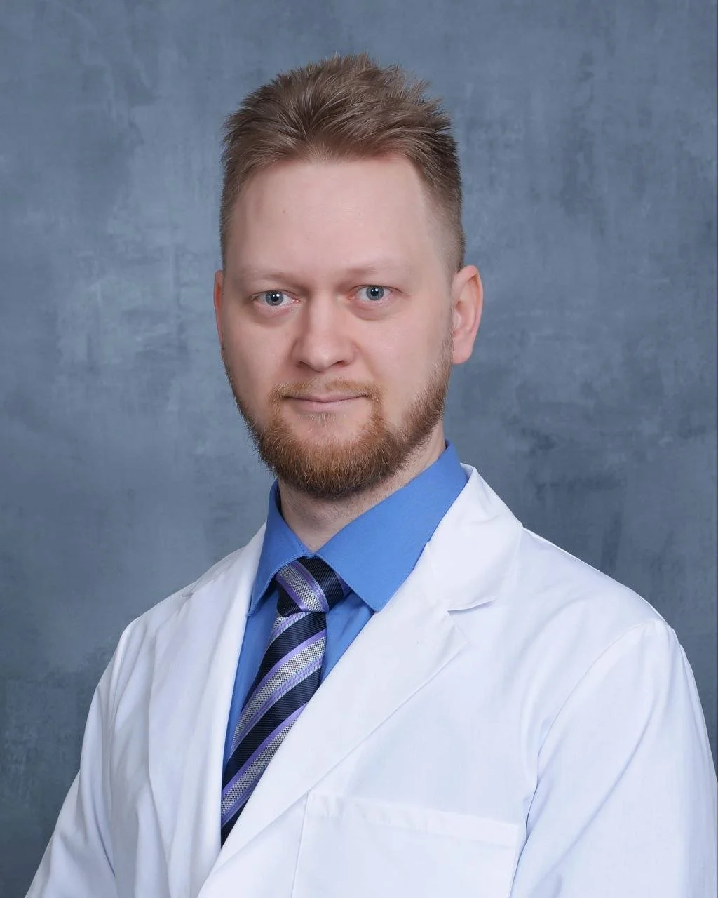 A man with light brown hair, a beard, and blue eyes wearing a white doctor coat, a blue dress shirt, and a purple striped tie, against a gray background.