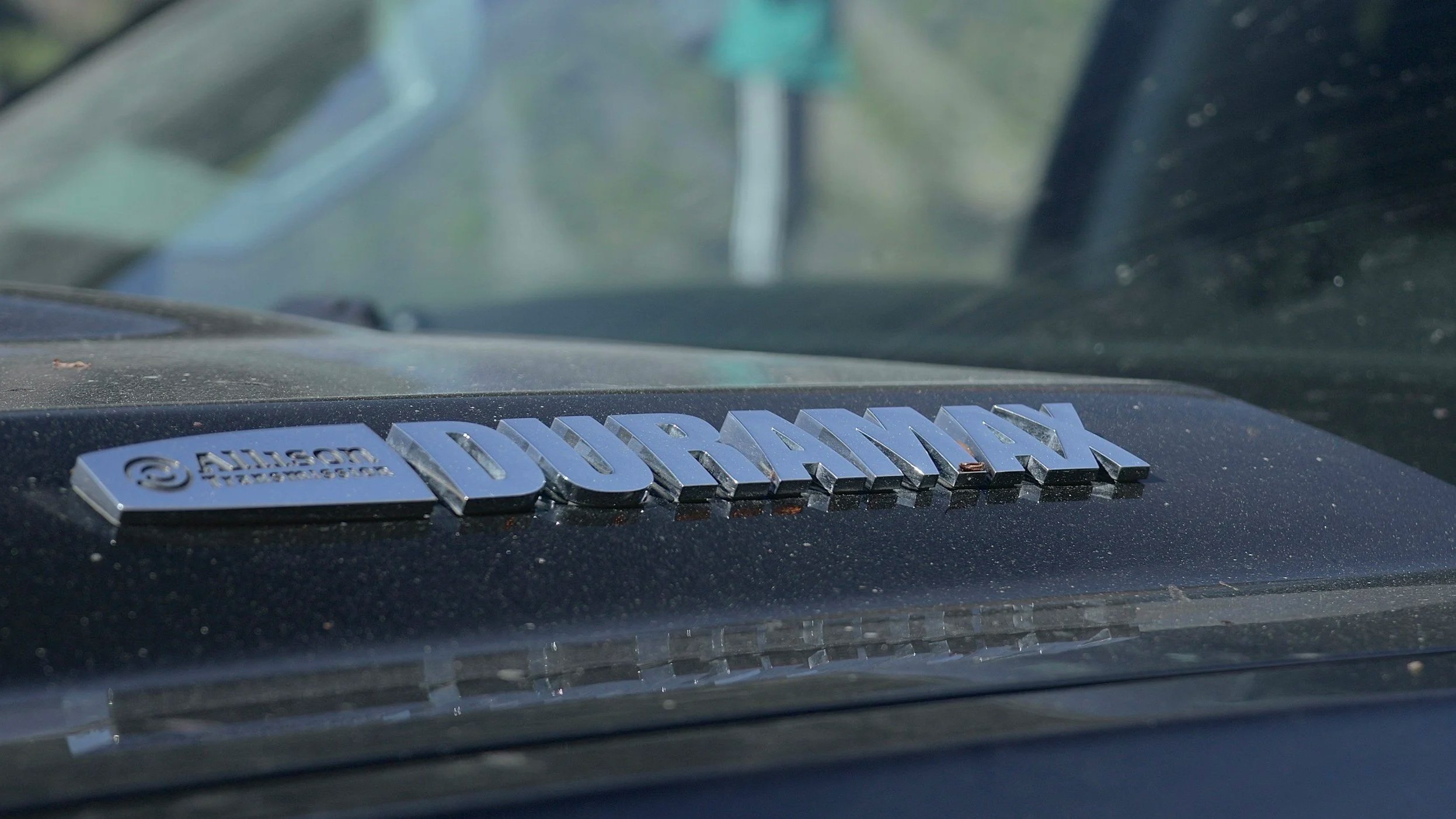 Close-up of a vehicle's badge on the dashboard that reads 'Duramax' with Allison Transmission logo beside it, on a dusty black surface with a blurred windshield and outdoor scenery in the background.