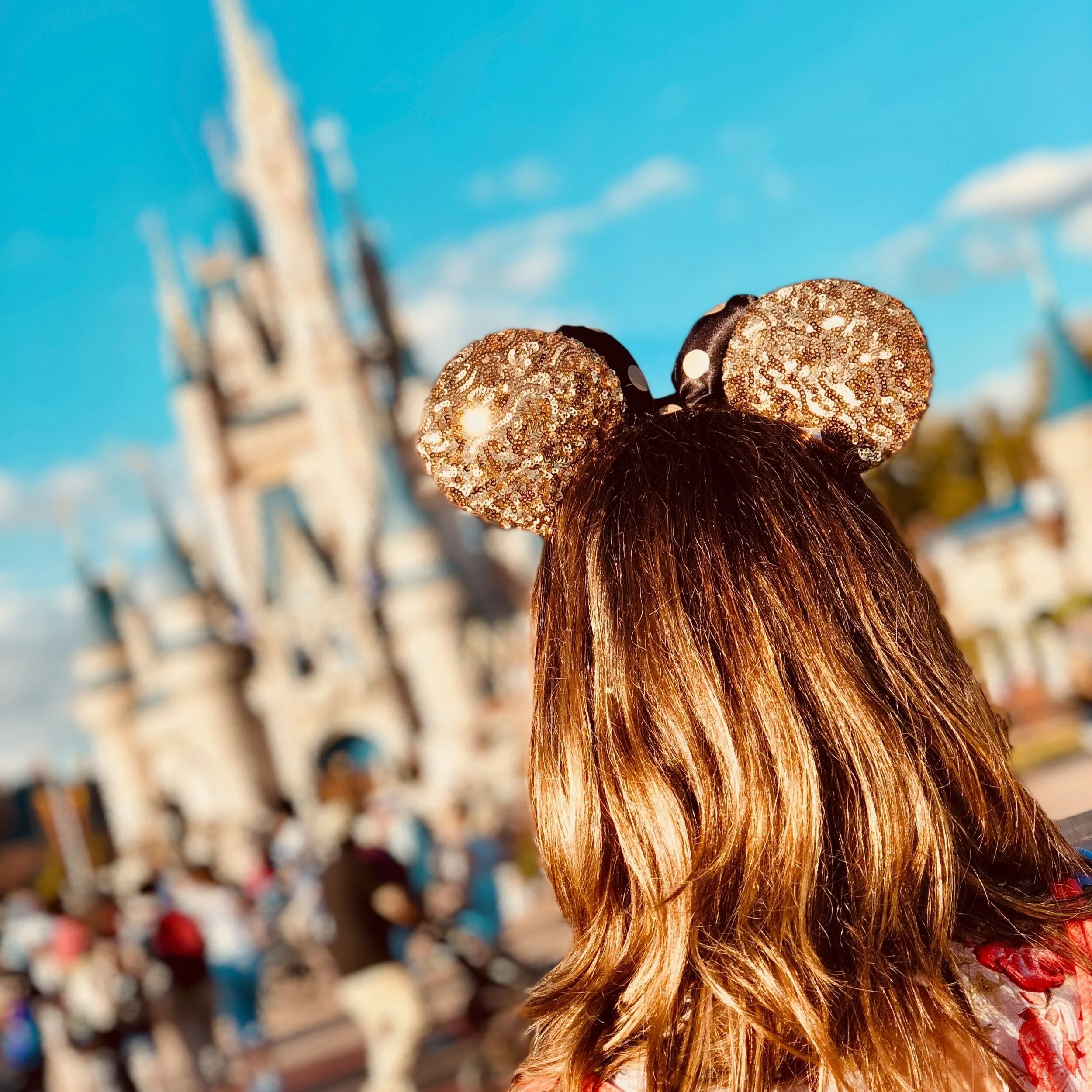A person with long, wavy, brown hair wearing Mickey Mouse ears with a glittery gold design, standing in front of the Disney castle at a theme park.