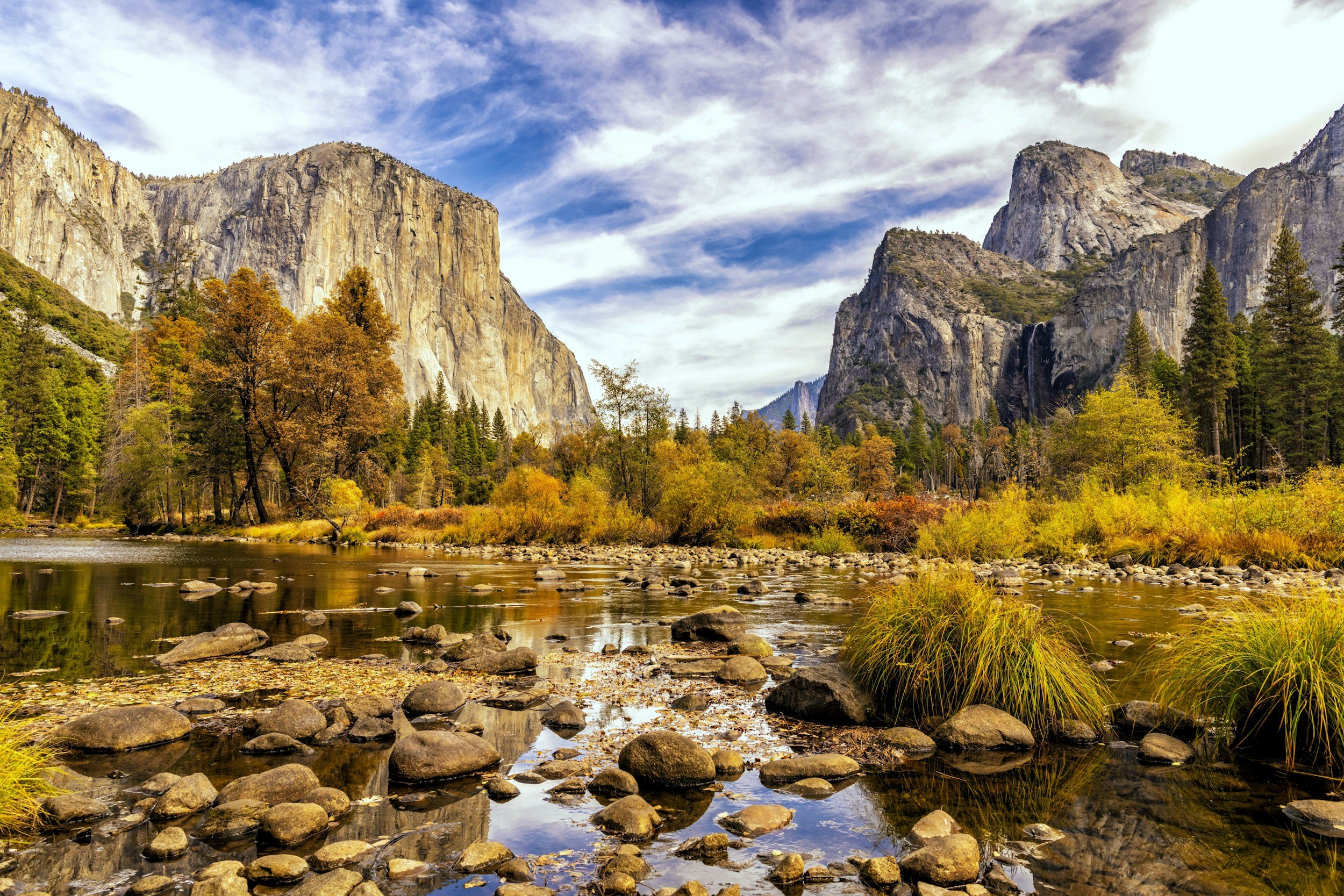 A scenic landscape of Yosemite National Park featuring a river with rocks in the foreground, surrounded by colorful autumn trees, with granite cliffs and waterfalls in the background under a partly cloudy sky.