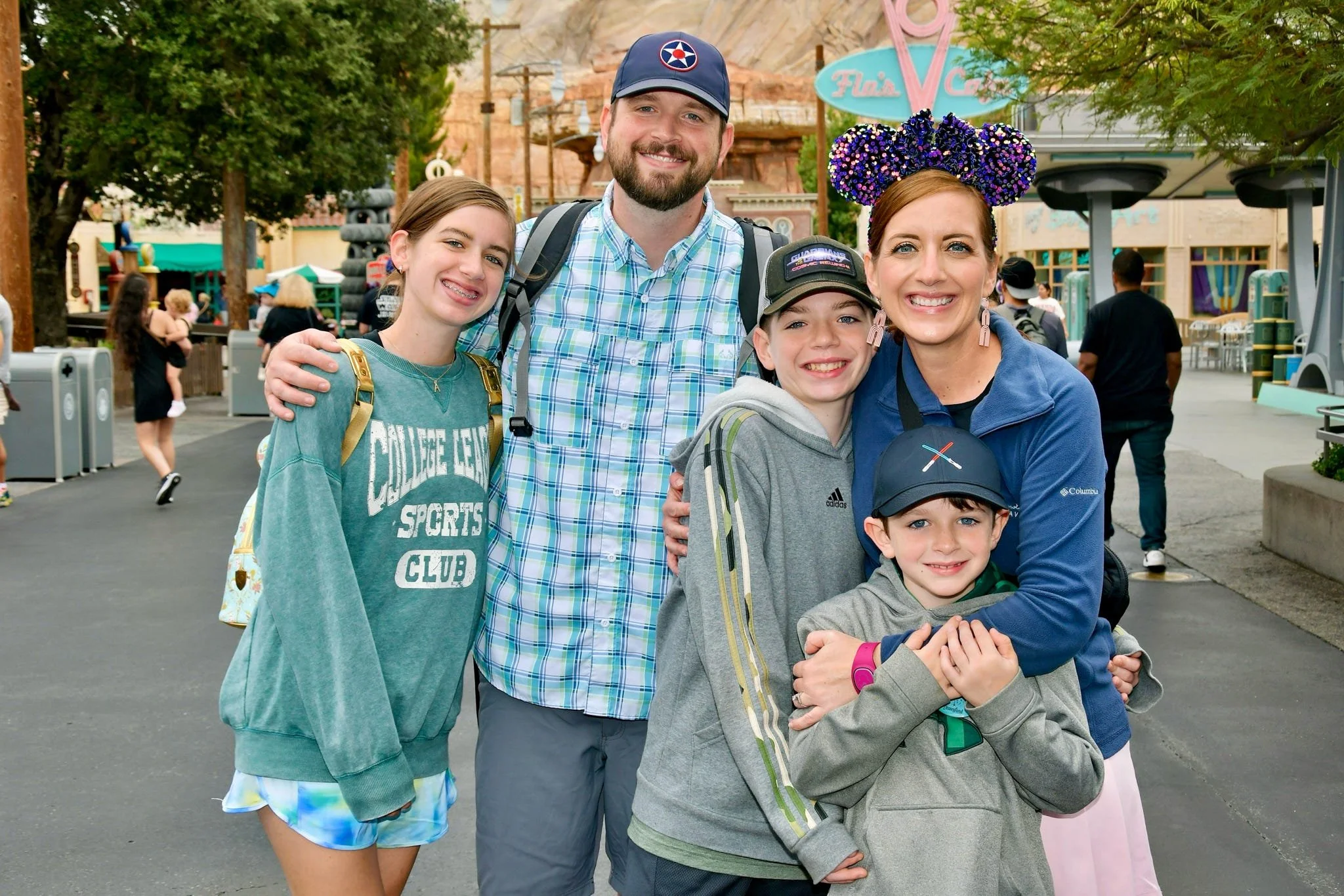 A smiling family of six posing for a photo at an amusement park with colorful attractions and trees in the background.