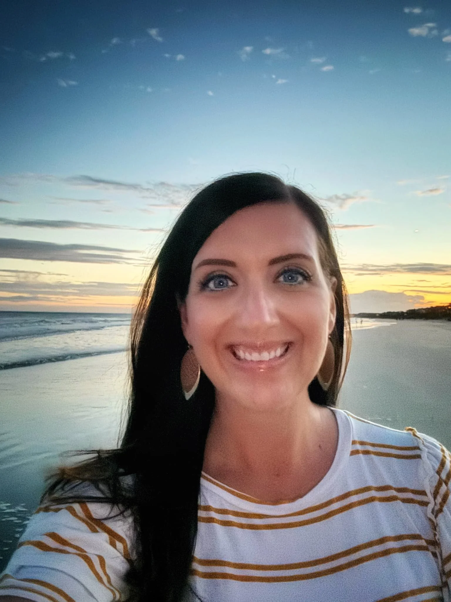 A woman with dark hair and blue eyes smiling on the beach during sunset, wearing a white shirt with gold stripes and large earrings.