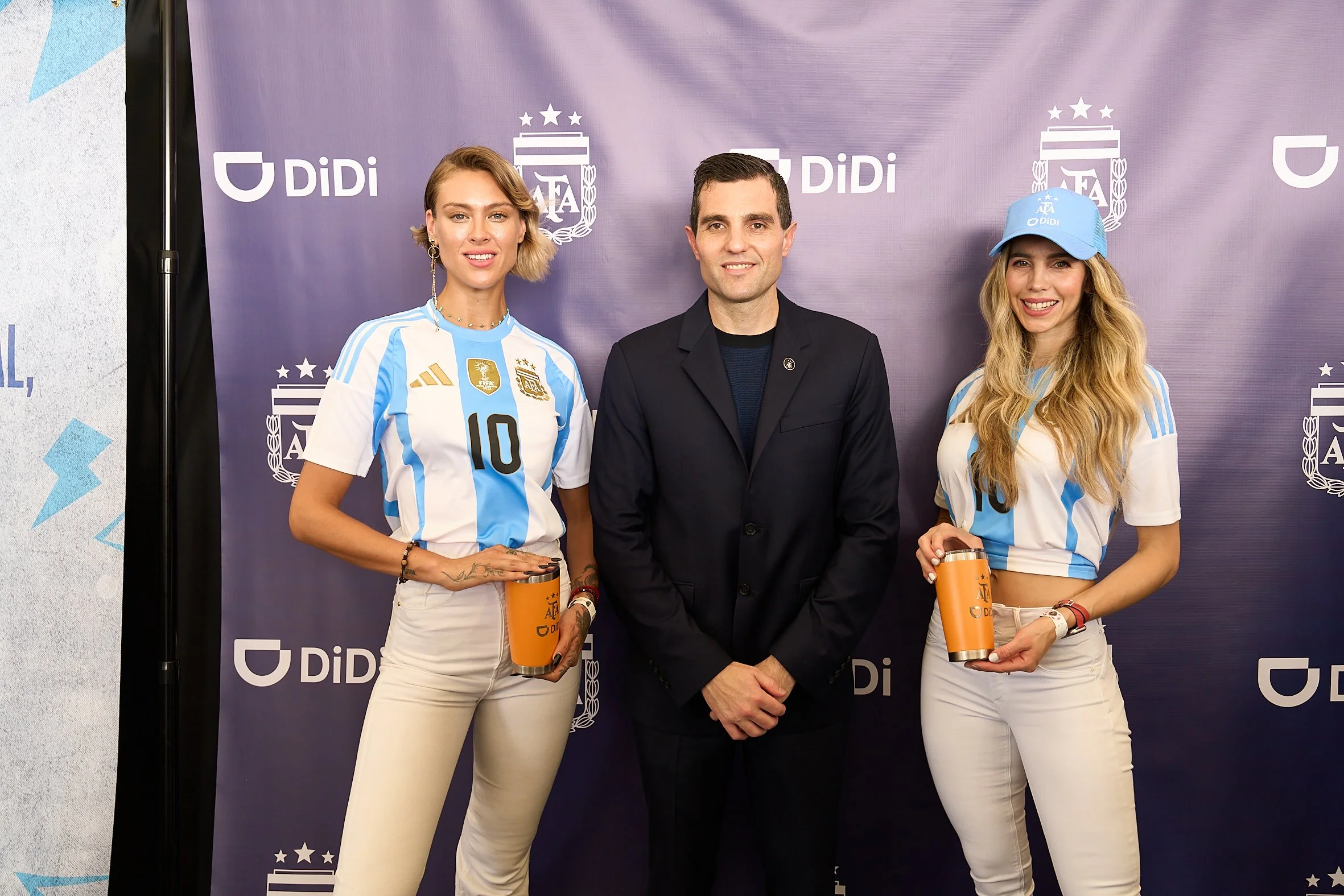 Two women wearing Argentina national soccer team jerseys and a man in a dark suit standing in front of a purple backdrop with 'Didi' and Argentina Football Association logos. The women are holding orange tumblers.