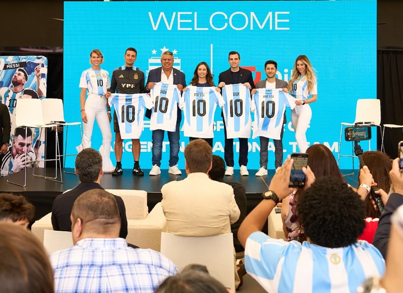 Group of six people on stage, holding up Argentina national football team jerseys with Messi's name and number 10, at a welcoming event with a large blue screen behind them displaying the word 'WELCOME' and the Argentina crest.