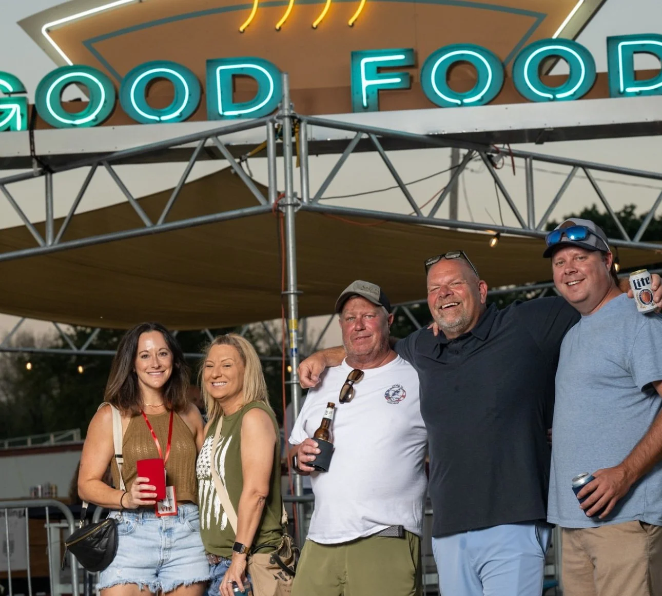 Group of five people smiling outdoors at a social gathering in front of a neon sign that reads 'Good Food,' with some holding drinks, during sunset or early evening.