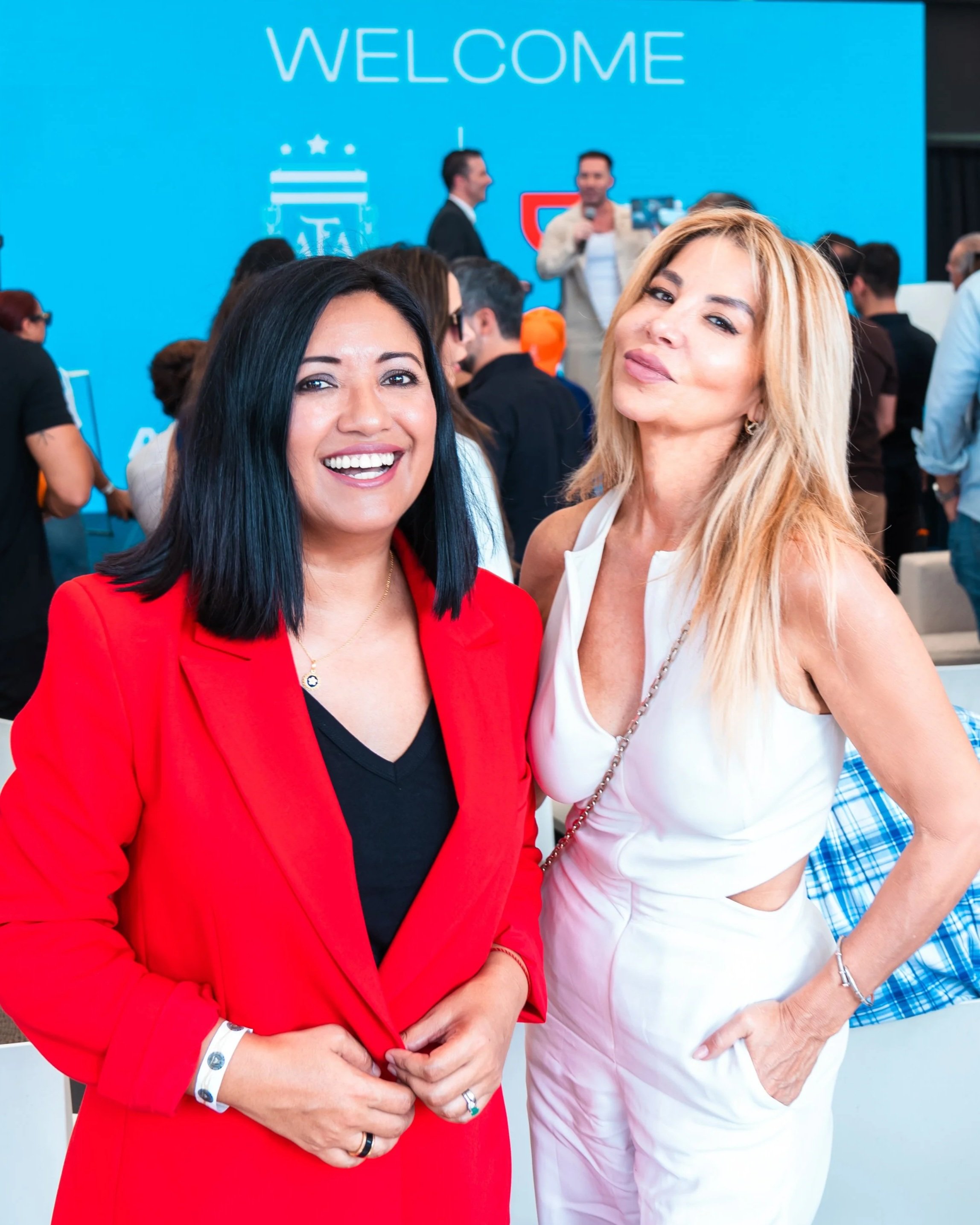 Two women smiling at a conference or event with a large blue welcome sign in the background. The woman on the left wears a red blazer and black top, the woman on the right wears a white sleeveless outfit.