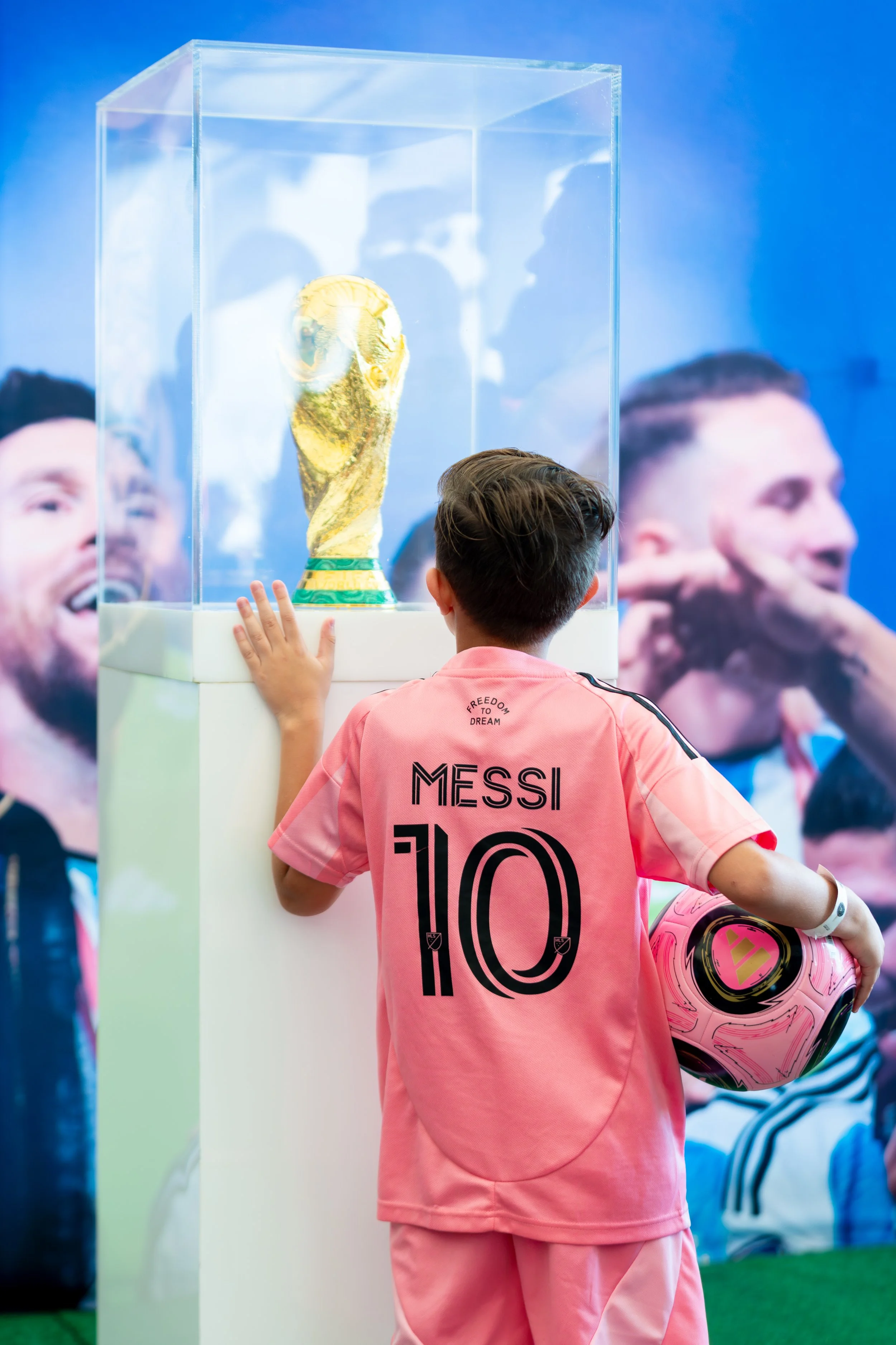 A young boy wearing a pink Messi jersey and holding a pink soccer ball looks at the FIFA World Cup trophy inside a transparent display case.