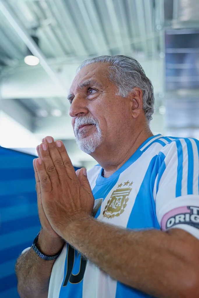 An older man in an Argentina national soccer team jersey with folded hands in a praying or greeting gesture, looking thoughtfully to the side in an airport or modern indoor setting.