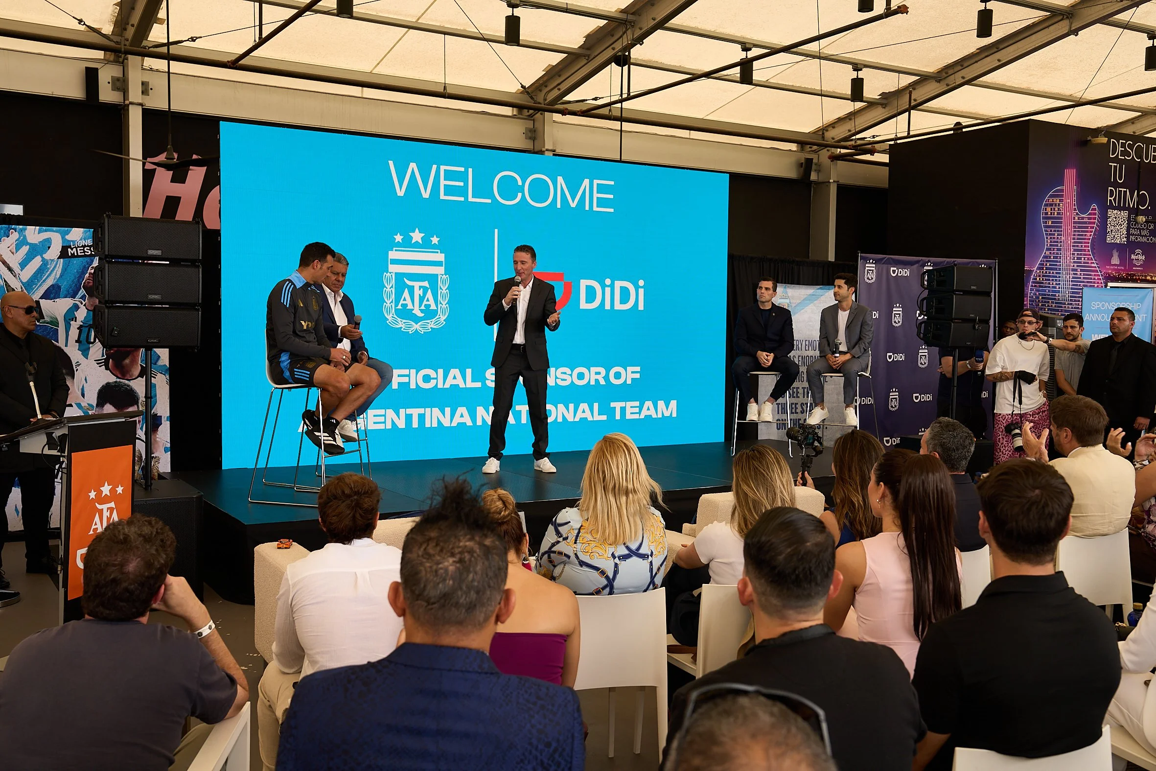 A large indoor event with a stage featuring a blue screen that reads 'Welcome' and includes logos of Argentina and Didi, with a speaker in a suit addressing an audience and several panelists seated on stage. Audience members are seated facing the sta