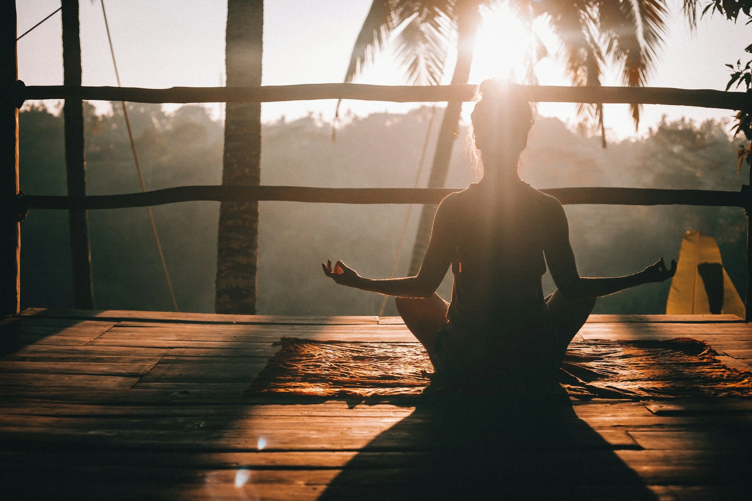 A woman practicing yoga in a cross-legged seated meditation pose on a mat during sunrise or sunset on a wooden deck outdoors, with palm trees and a tropical landscape in the background.