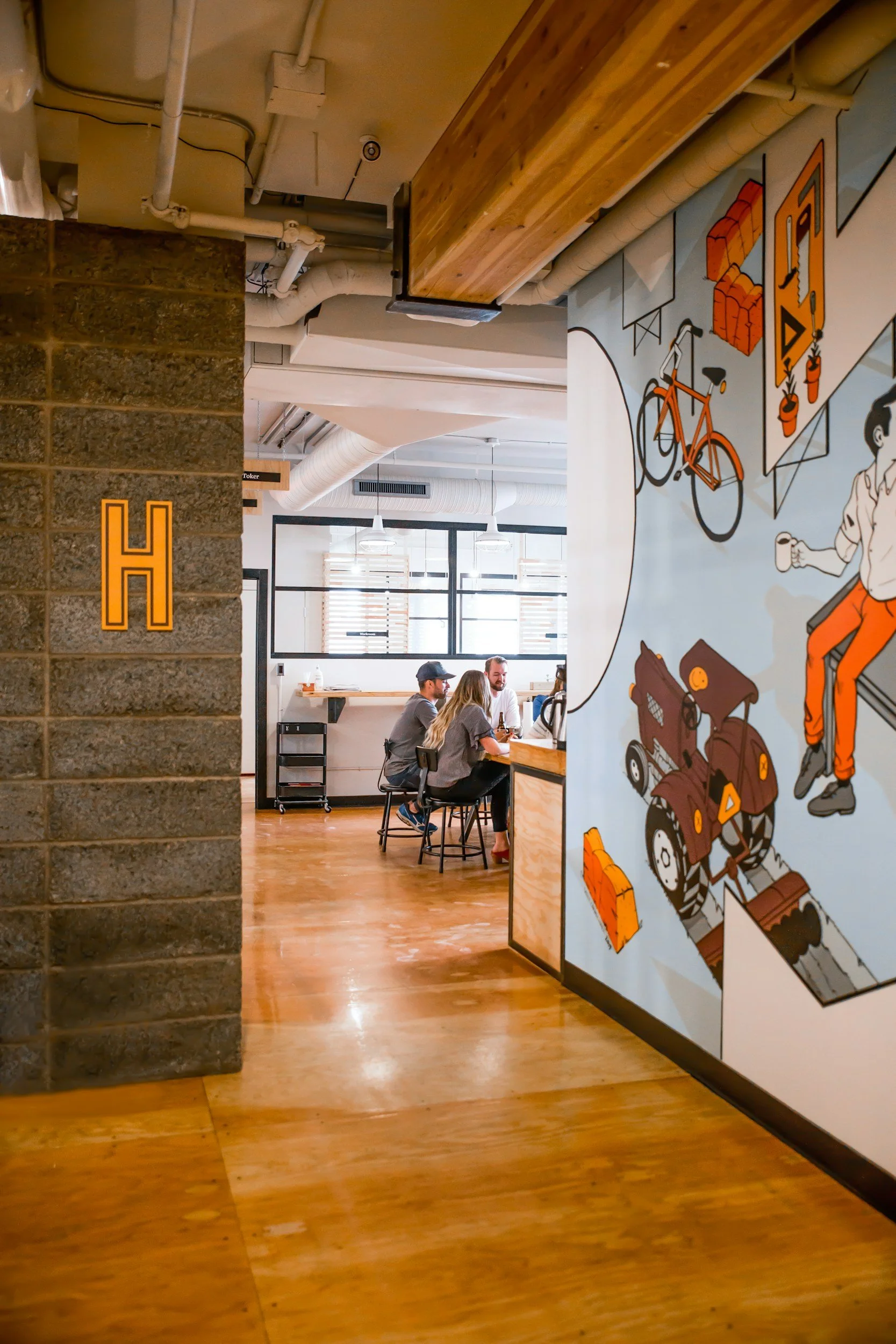 Interior of a modern cafe with wooden flooring, a brick wall with an orange letter H, and a colorful mural with bicycles, a tractor, and a person sitting and drinking coffee.