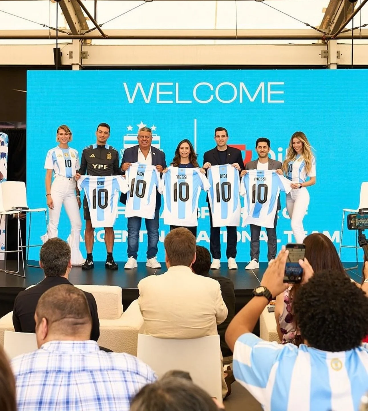 Group of people standing on stage at a welcome event for an Argentine national football team, holding up Argentine soccer jerseys with the number 10. Audience members are taking photos.