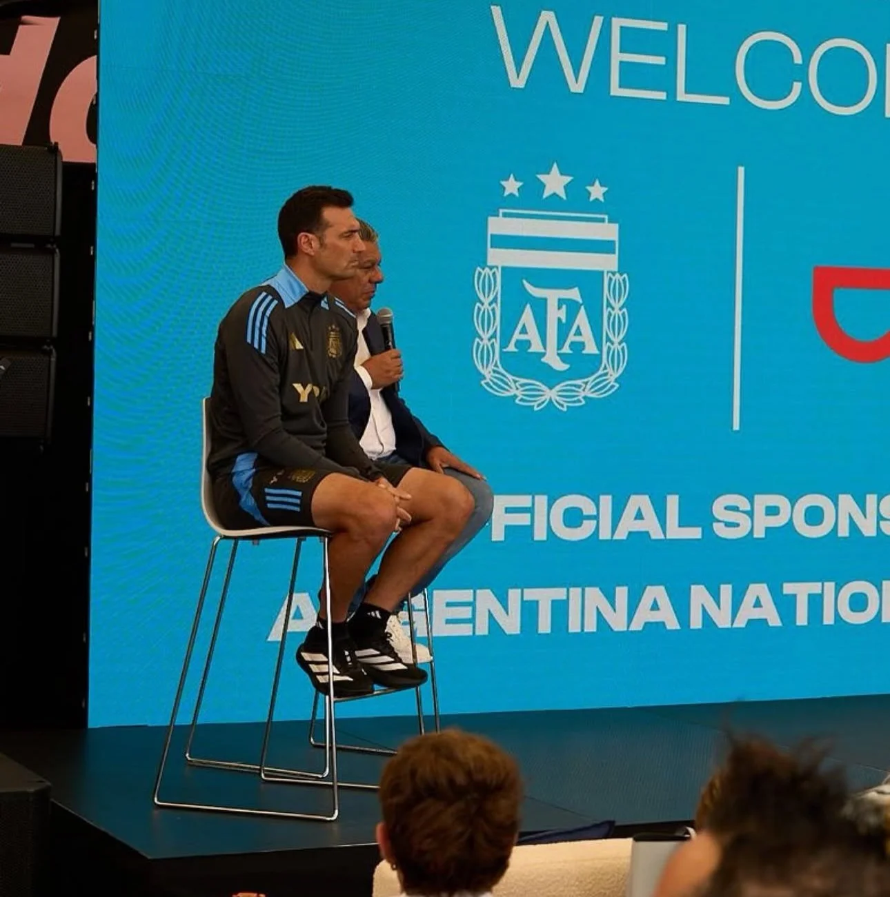 A man in a sports uniform sitting on a stool on stage, with a person next to him holding a microphone. The background displays a large blue screen with the Argentine Football Association (AFA) logo and the words "Welcome" and "Official Sponsors Argen