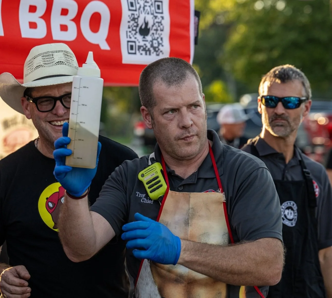 Three men outdoors, one holding a large clear bottle with a yellow liquid, wearing a cowboy hat and glasses, another in an apron with a walkie-talkie, and the third in sunglasses in the background, at a barbecue event.