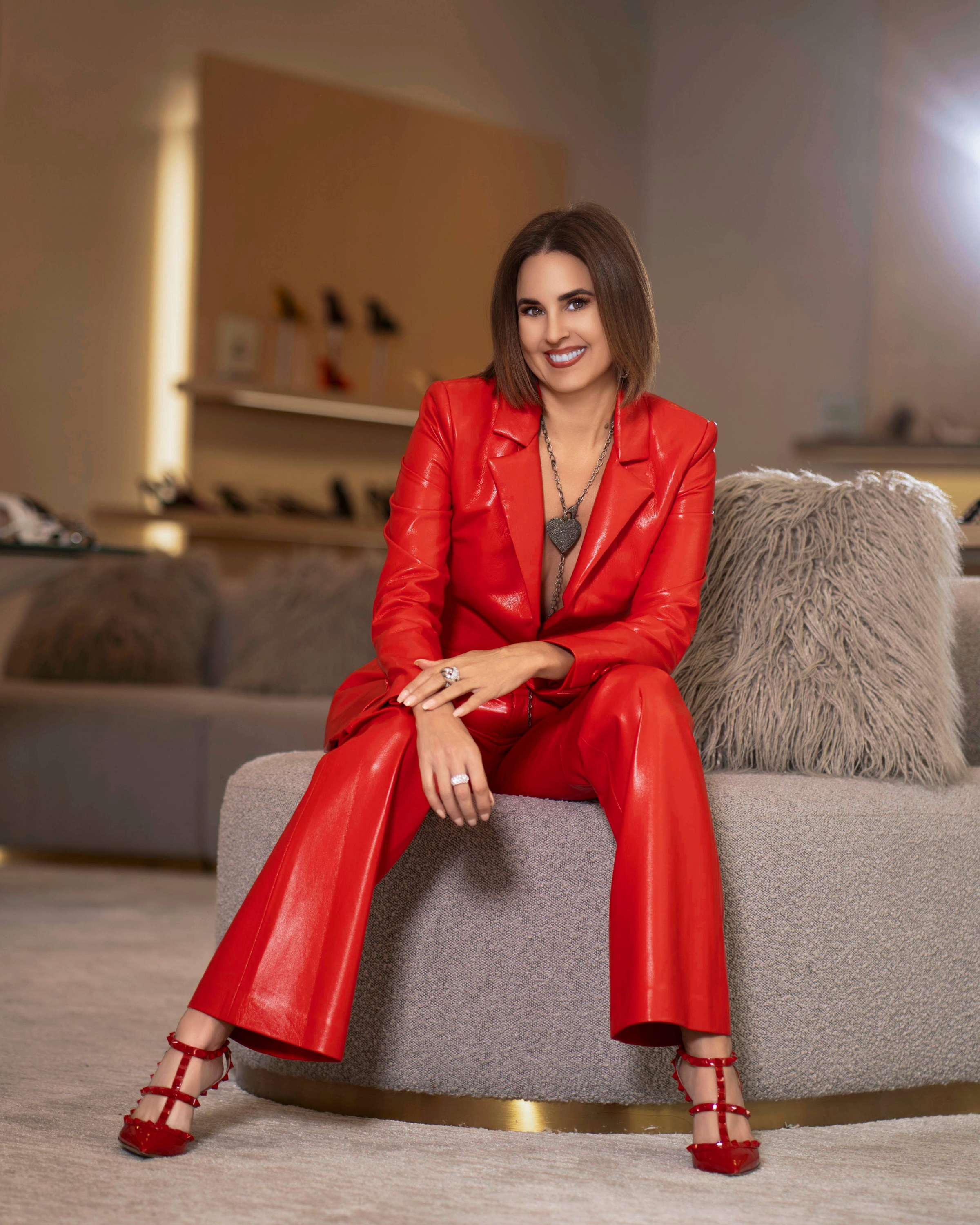 A woman with shoulder-length brown hair and makeup, sitting on a gray round sofa in a living room, wearing a red leather suit and red high heels, smiling at the camera.