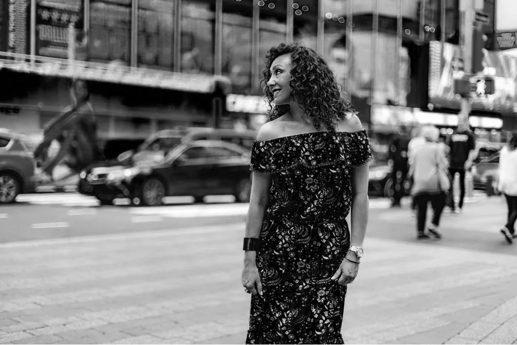 A woman with curly hair smiling and walking on a busy city street in black and white.