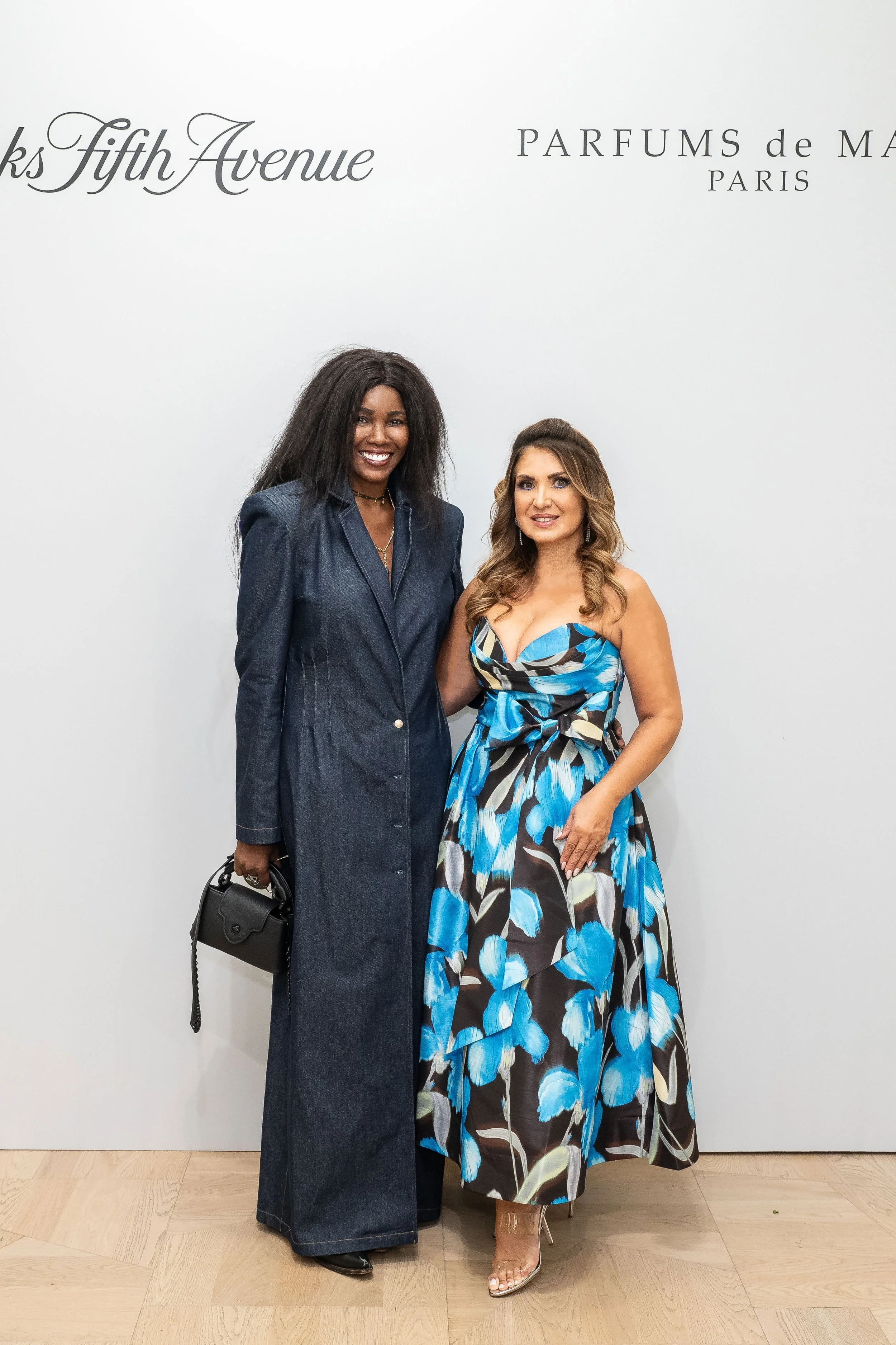 Two women standing side by side in front of a white wall with text that reads 'Books Fifth Avenue' and 'PARFUMS de MARC PARIS.' The woman on the left is wearing a dark denim jumpsuit and holding a black handbag, while the woman on the right is wearing a blue and black floral sleeveless dress with a bow at the waist and clear high heels.