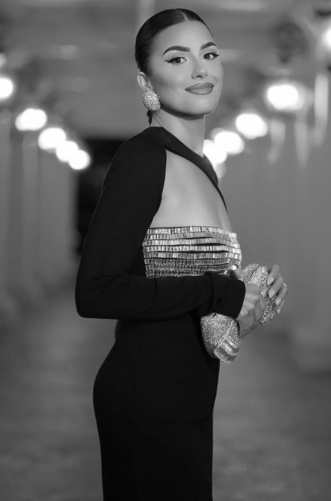 A woman with dark hair in a sleek bun wearing large earrings, a fitted dress with a shimmering strip, and holding a glittering clutch purse standing in a hallway with lights in the background.
