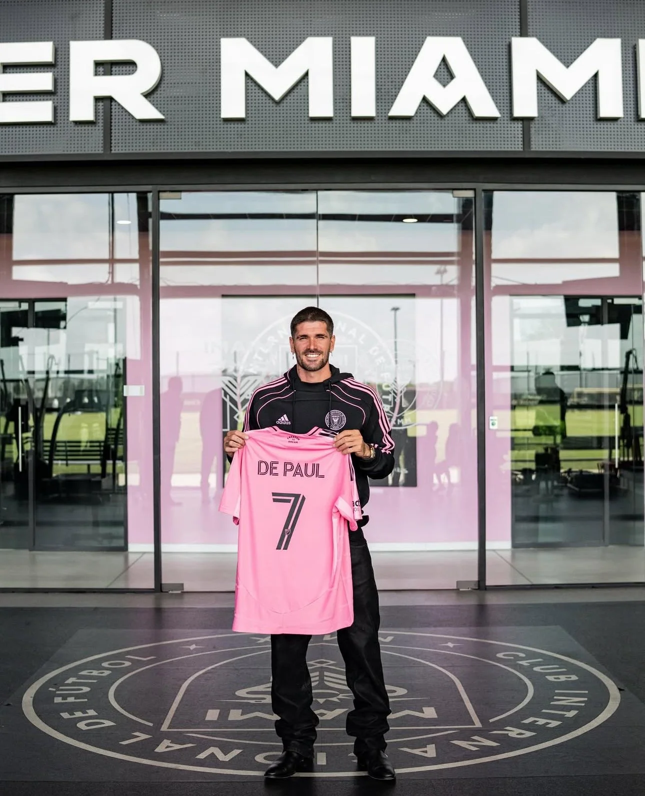 A man standing inside a building holding a pink soccer jersey with the name 'DE PAUL' and the number 7. The building has large glass doors and a sign above with the text 'FIER MIAMI.'