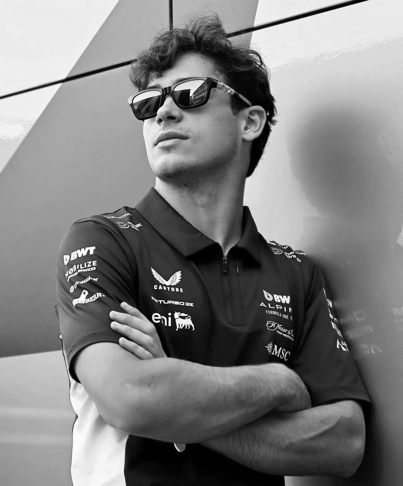 A young man wearing sunglasses and a racing team shirt with various sponsor logos stands with his arms crossed, looking to the side, against a geometric background.
