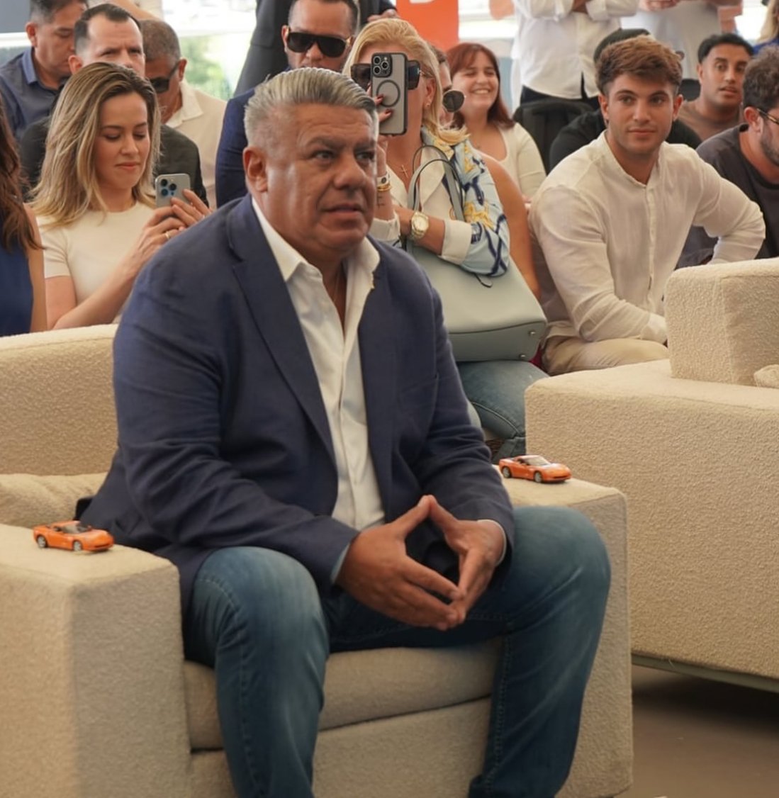 Man in a navy blazer and white shirt sitting on a beige couch, surrounded by people in an indoor setting, with some taking photos and videos.