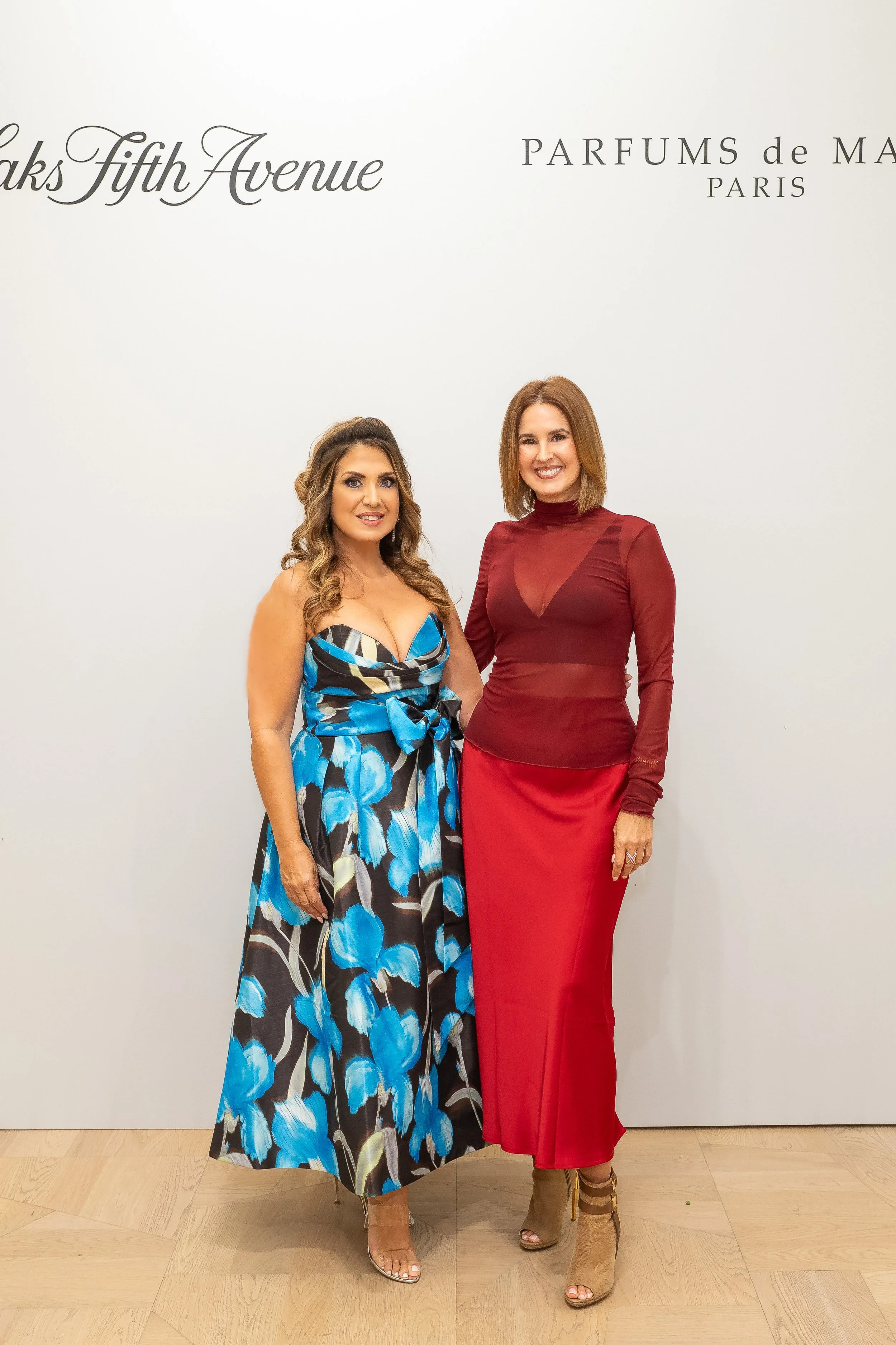 Two women standing and smiling in front of a white wall with black text. The woman on the left wears a sleeveless, floral dress in black, blue, and white with a large bow at the waist, and beige high-heeled sandals. The woman on the right wears a long-sleeved, burgundy sheer top, a red satin skirt, and beige high-heeled ankle boots.