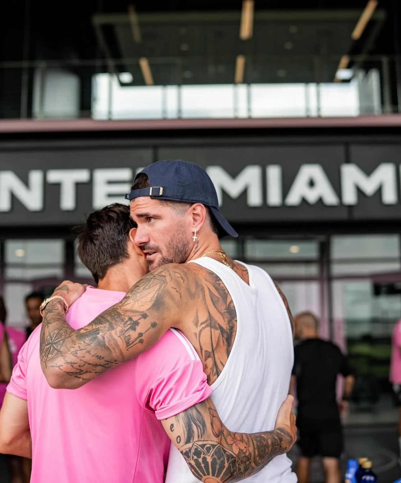 Two men hugging, one with tattoos, wearing a navy baseball cap backward, a cross earring, and a chain; the other with short dark hair, wearing a pink shirt. They are at an airport with a sign that reads 'MIAMI' in the background.