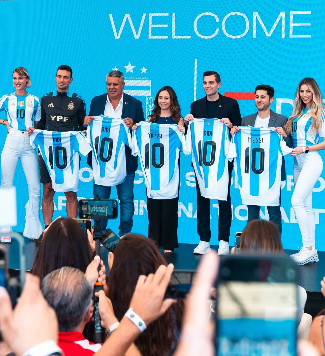Group of seven people holding Argentina soccer jerseys with the name Messi and the number 10, standing in front of a blue background that says "Welcome" and some symbols, with a crowd taking pictures in the foreground.