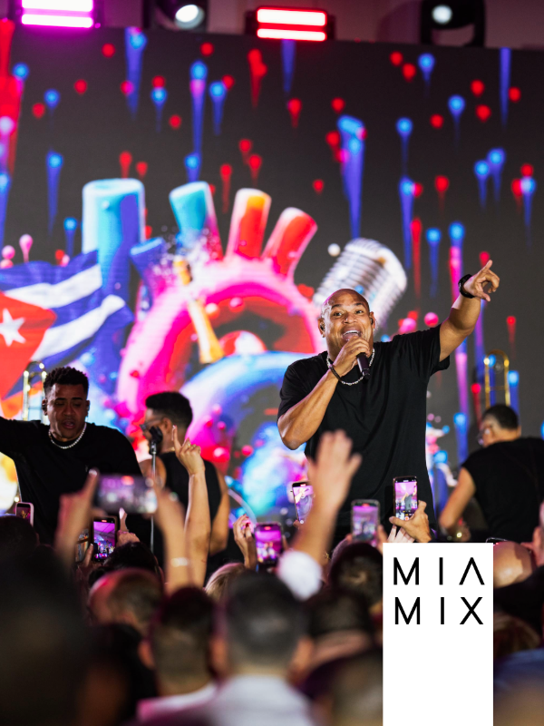 A live concert with a male singer on stage, holding a microphone and pointing, surrounded by backup singers, with a crowd taking photos, and a vibrant, colorful digital backdrop with neon and abstract shapes.