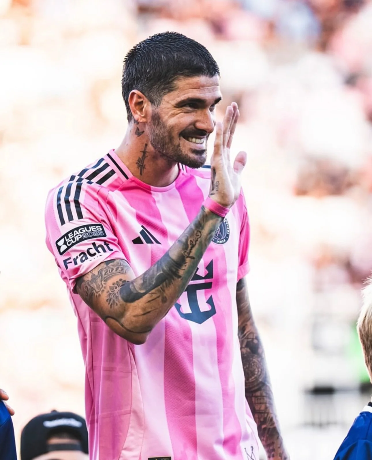 A male soccer player with tattoos on his arms and neck, wearing a pink jersey with black stripes on the shoulders, smiling and waving.