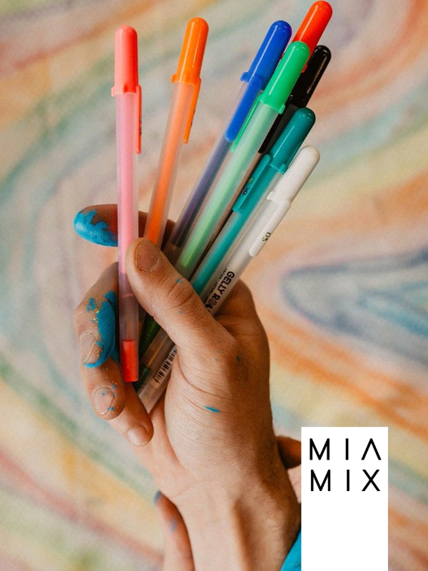 A hand holding a bunch of colorful markers with blue paint on fingers and thumb, against a blurred, multicolored background.
