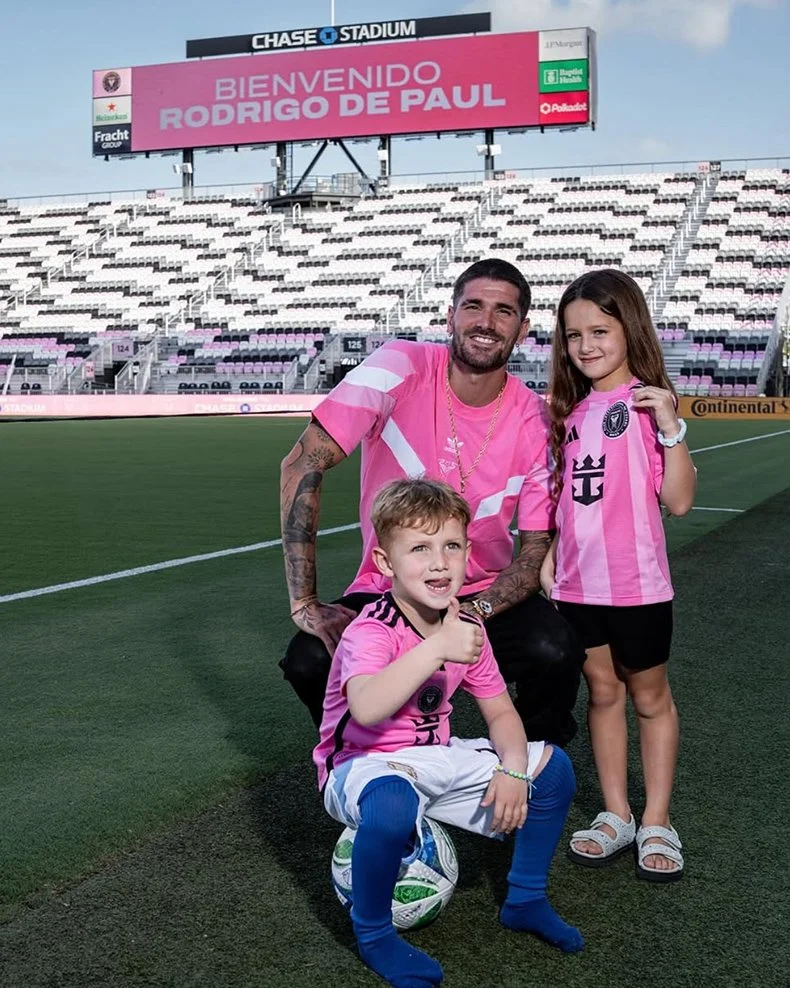A man and two children in pink soccer jerseys on a soccer field at Chase Stadium. The man is kneeling with both children, smiling, with a large welcome sign for Rodrigo De Paul in the background.