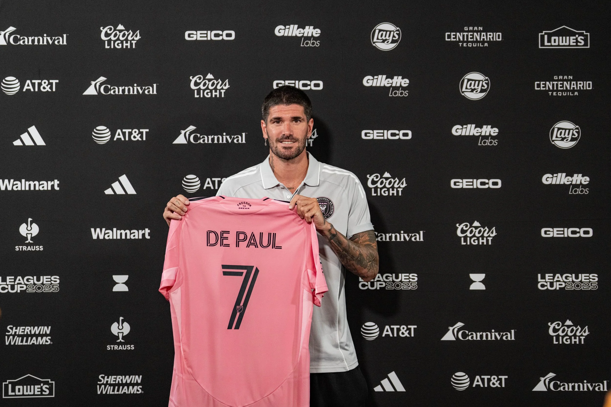 A man in a white shirt holding up a pink soccer jersey with the name 'De Paul' and the number 7 on it, standing in front of a backdrop with various sponsor logos.