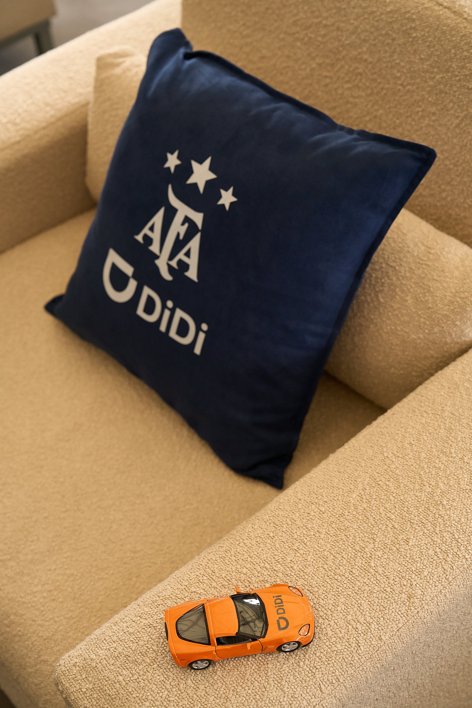 A beige couch with a navy blue pillow that has a design with stars and the text 'AFA DIDI'. On the armrest of the couch, there is an orange toy car with the text 'DIDi' on it.