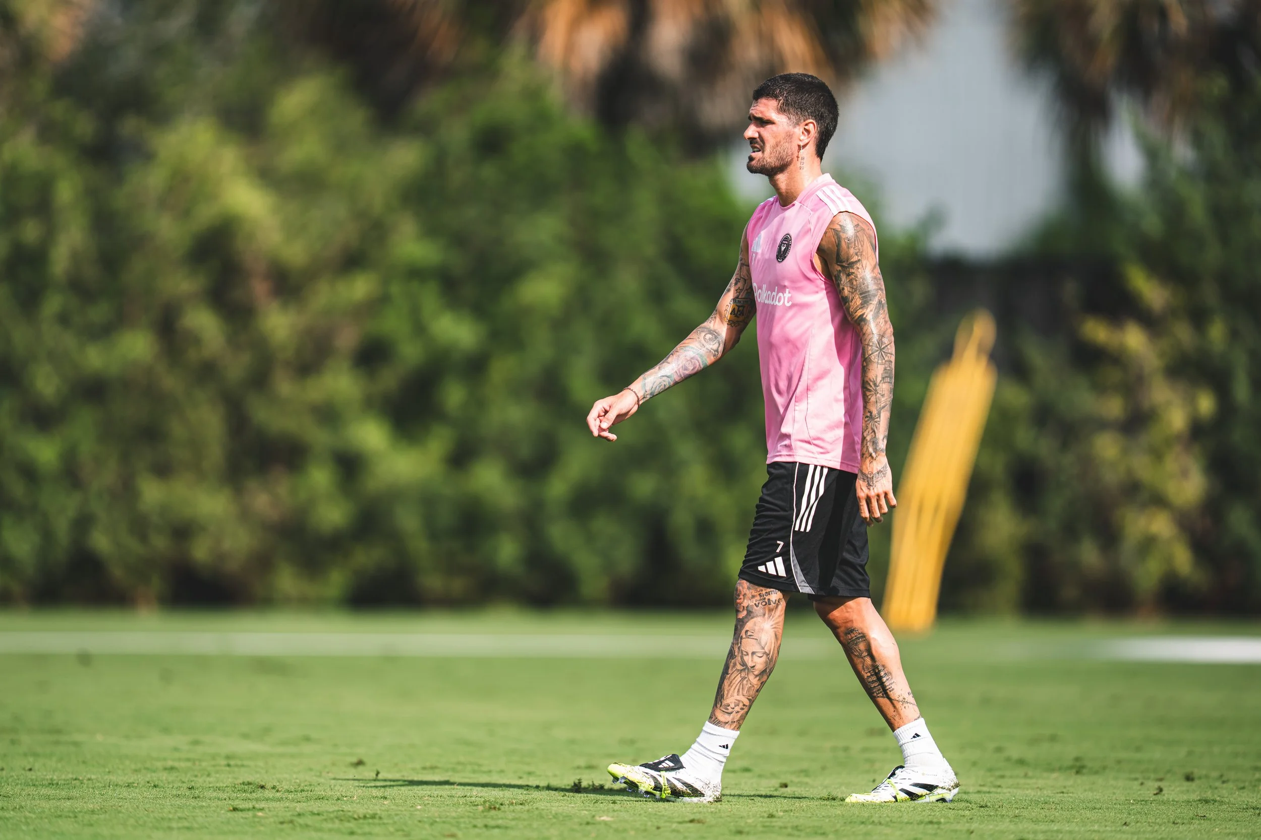 A tattooed man in a pink sports jersey and black shorts walking on a grassy field during daytime, with trees and a yellow training dummy in the background.