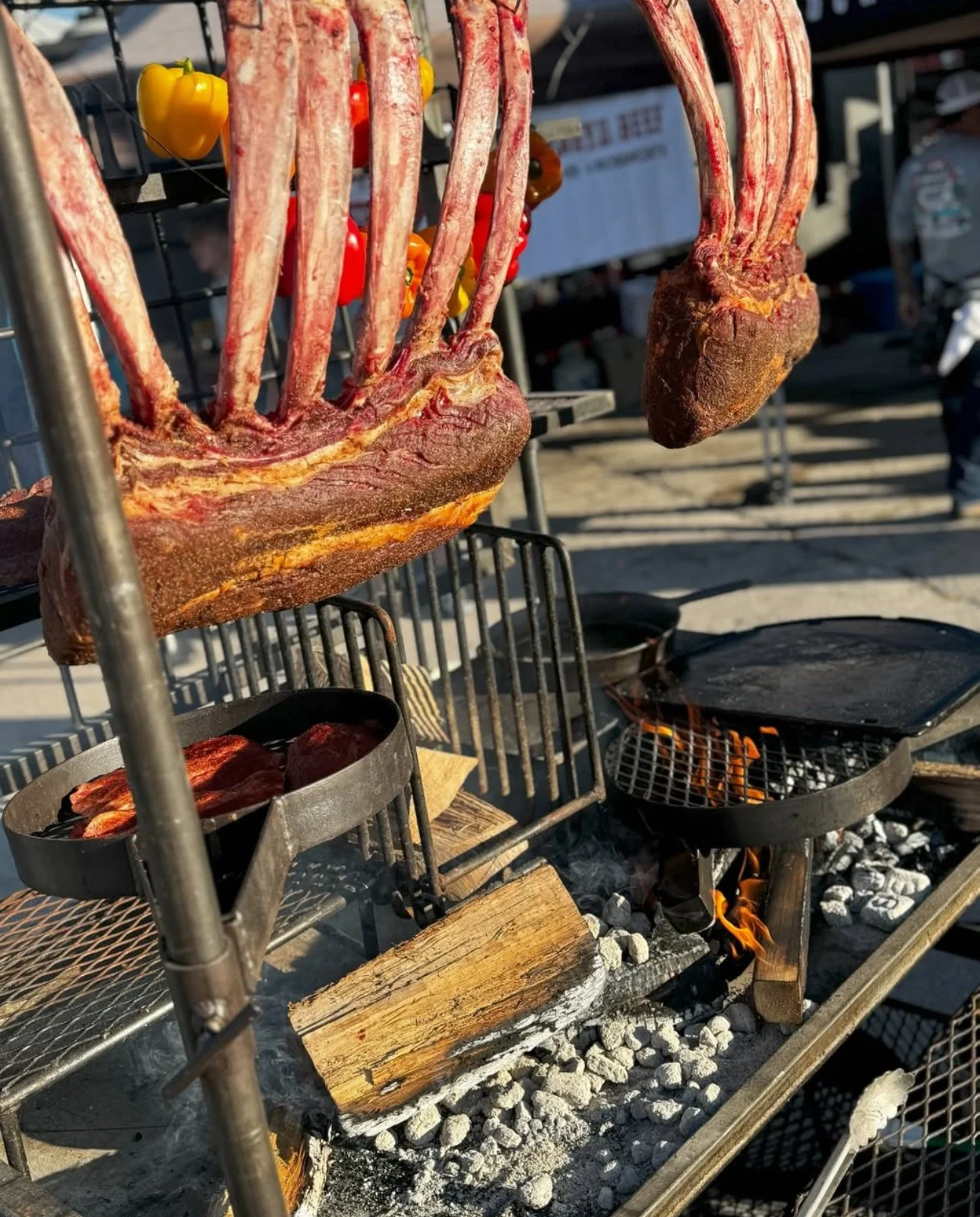 Lamb chops hanging over a charcoal grill with flames, on a metal rack with smoke and ashes.