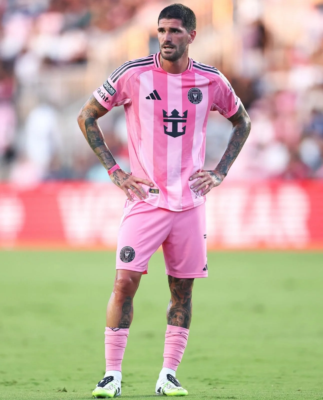 A male soccer player with tattoos, wearing a pink uniform with black and white accents, standing on the field with his hands on his hips, looking forward.
