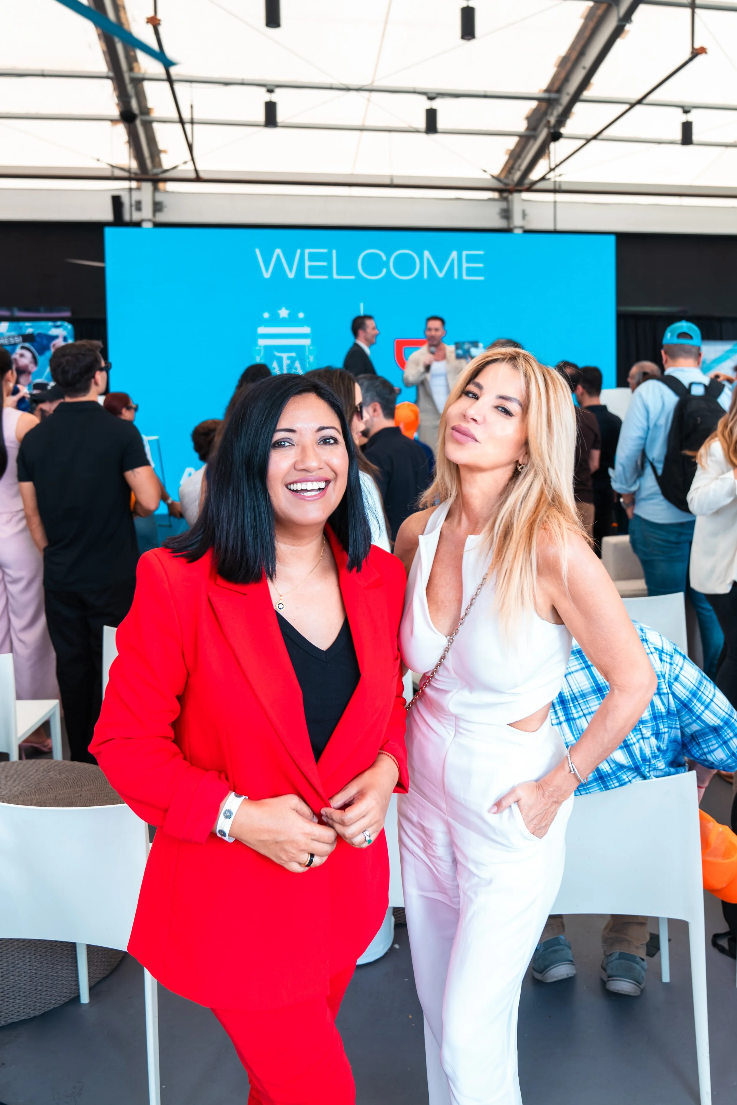 Two women smiling in front of a blue 'Welcome' sign at a crowded indoor event or conference.