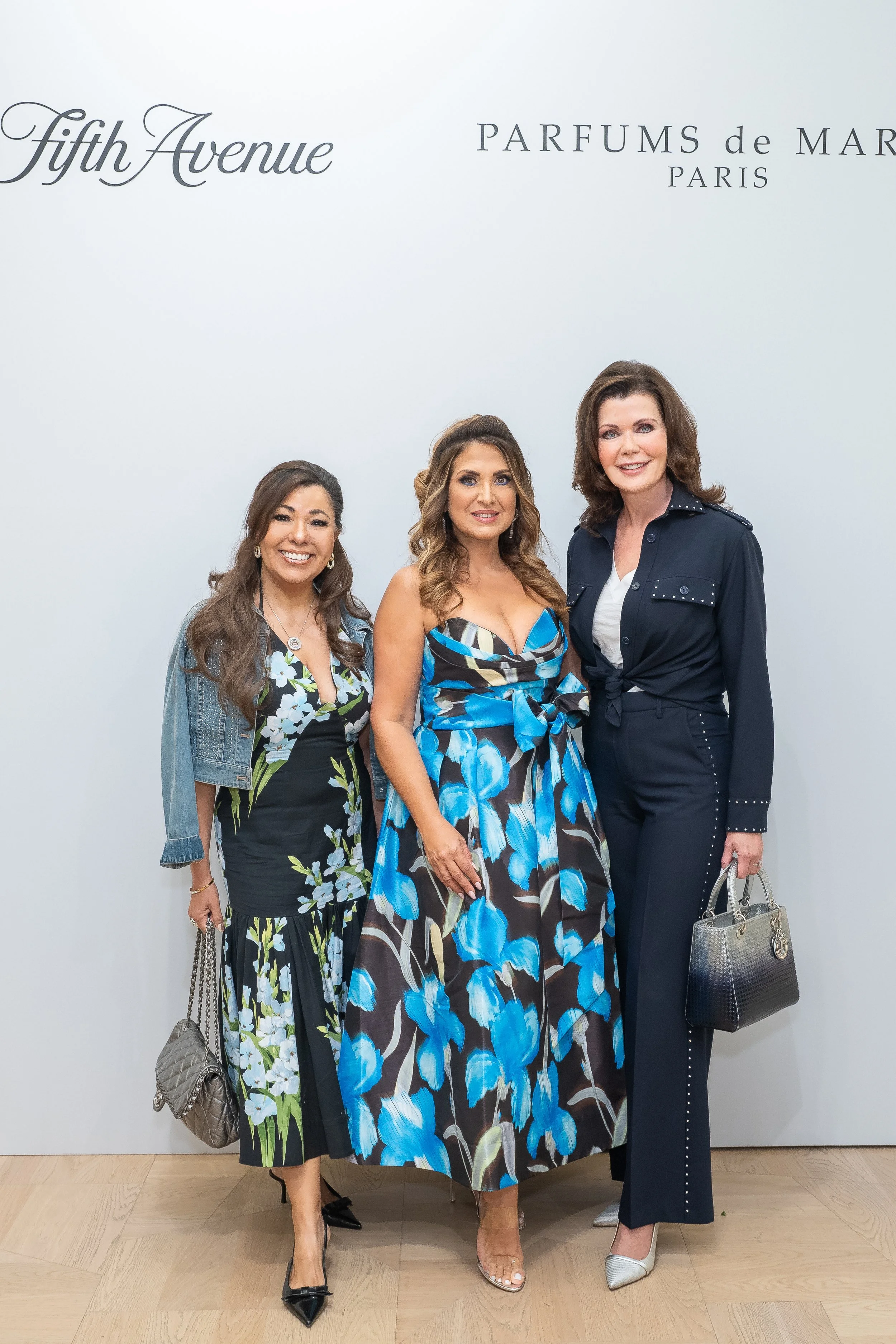 Three women standing together and smiling at a fashion event with a white background displaying the words 'Fifth Avenue' and 'Parfums de Marc Paris'.