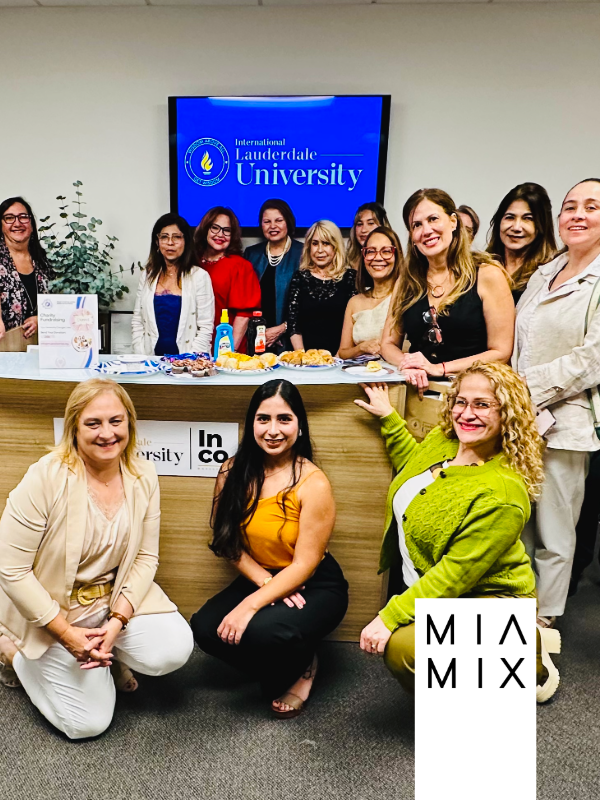 Group of women gathered around a table at International Lauderdale University, with a monitor displaying the university's logo in the background. The table has snacks and condiments.