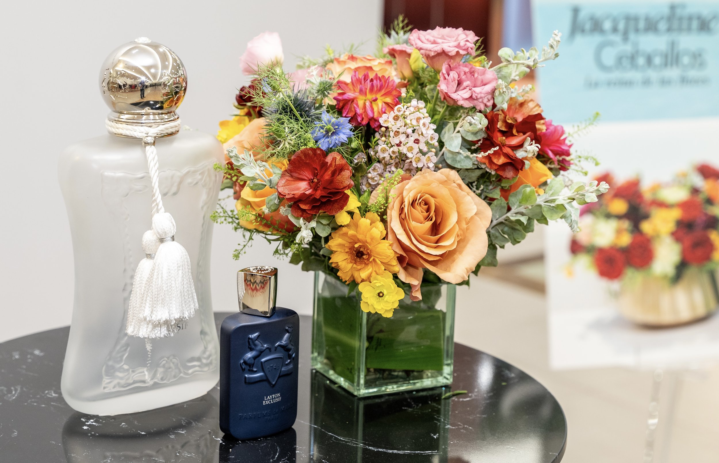 A still life arrangement featuring a colorful bouquet of flowers in a square glass vase and a decorative frosted glass bottle with a silver cap and a white tassel, placed on a black marble surface, with a bottle of perfume in front of the bottle and 