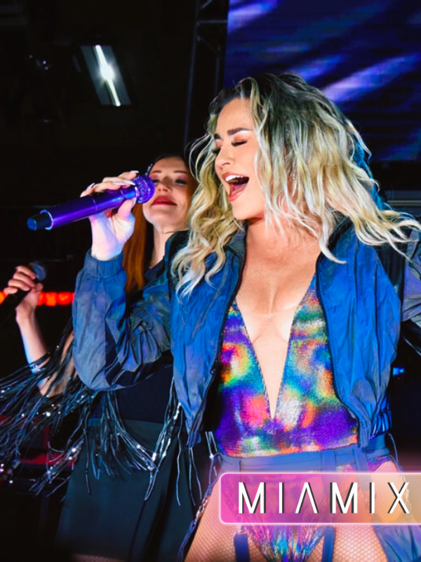 A woman with long, wavy blonde hair holding a red microphone to her mouth, wearing a metallic silver and striped colorful outfit with a black, shimmering jacket. She is on a stage or behind the scenes, with equipment visible in the background.