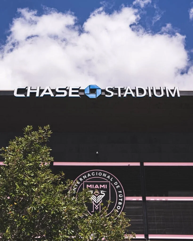 Exterior of Chase Stadium with the stadium name and logo at the top, partially obscured by a tree, under a partly cloudy sky.
