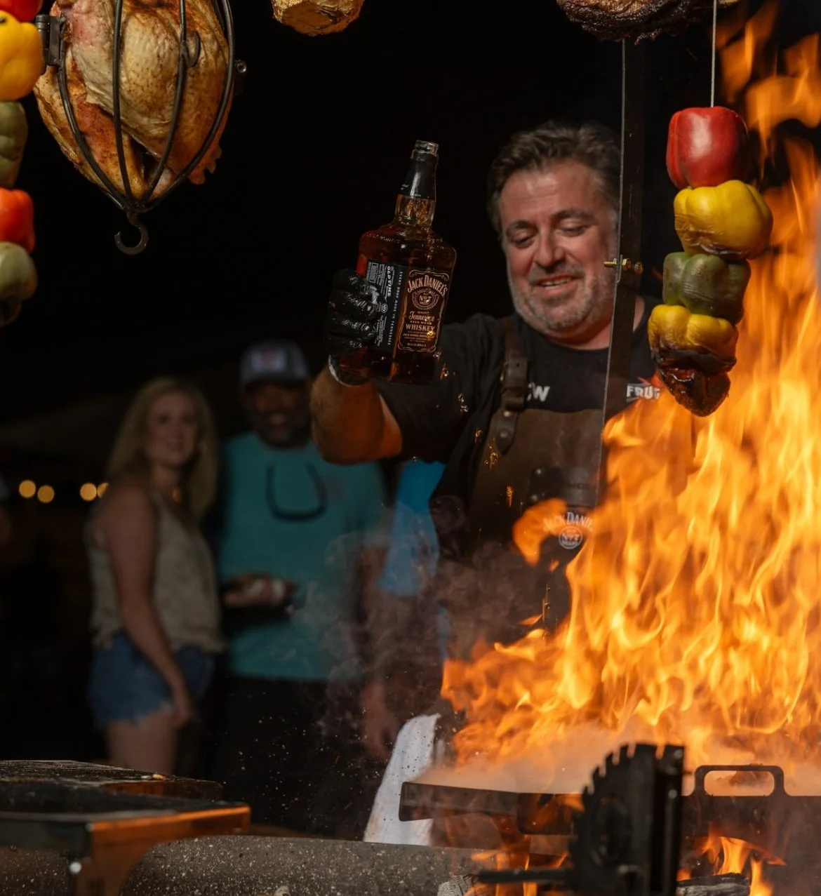 A chef holding a bottle of Jack Daniel's whiskey near a large flame with hanging skewers of colorful peppers and meats, while smiling at a lively outdoor event at night.