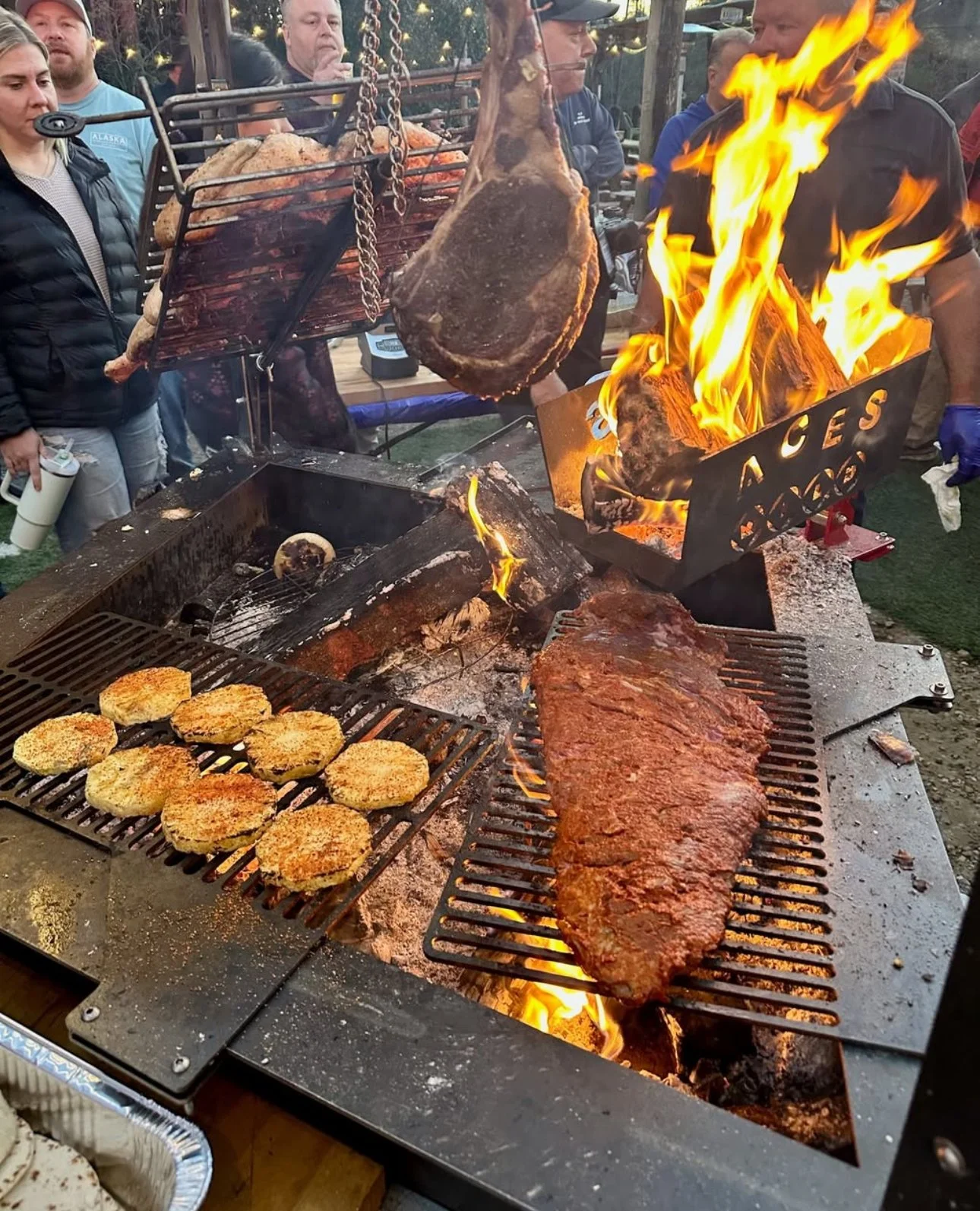 Outdoor barbecue scene with multiple people around a grill, grilling large cuts of meat, burgers, and breaded items, with flames and smoke evident.