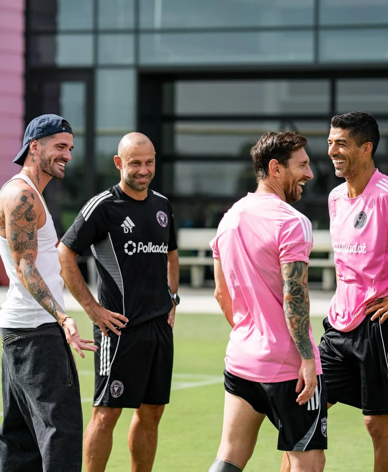 Four male soccer players on a field laughing and talking, one in a black uniform and the others in pink training shirts.