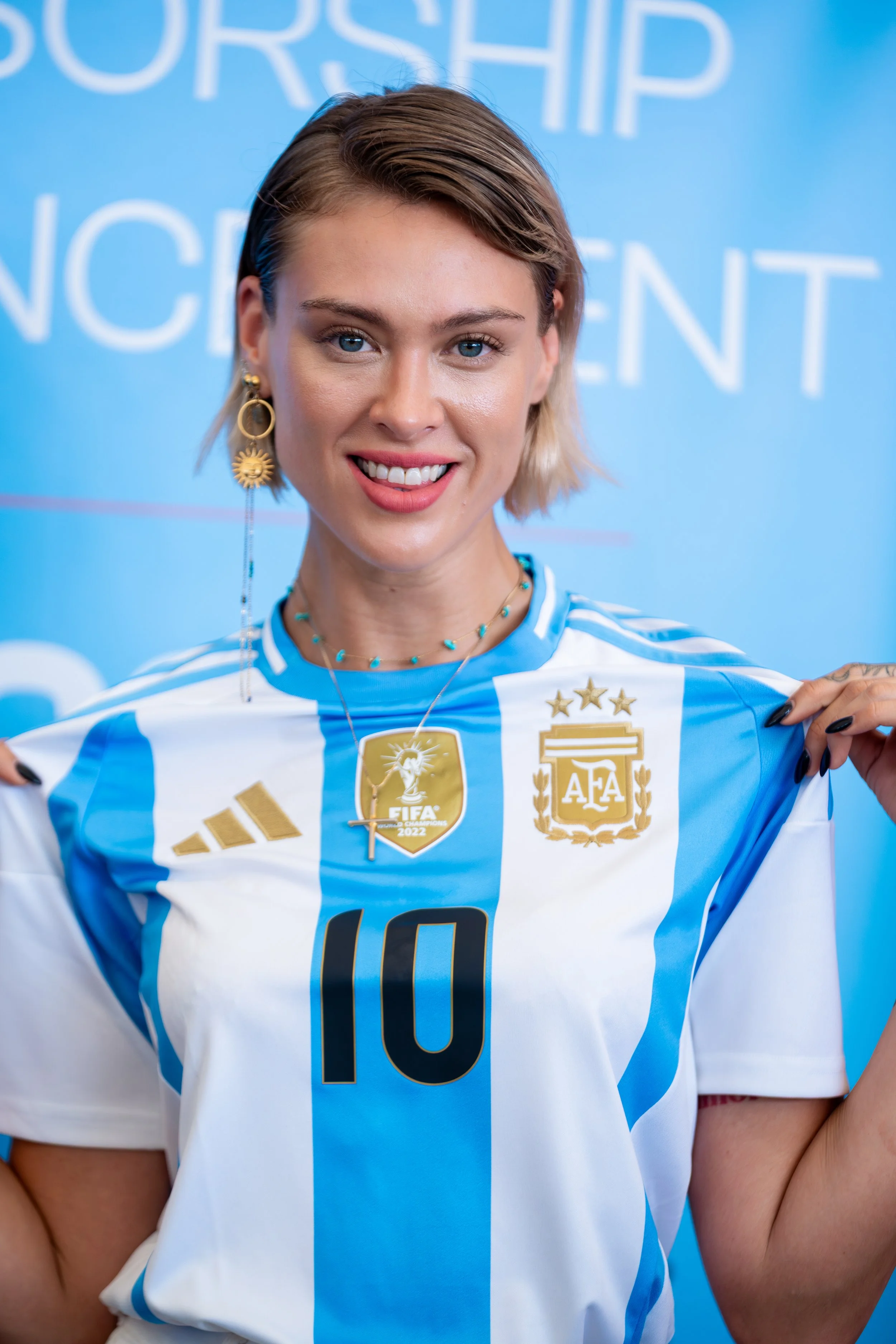 A woman smiling and wearing an Argentina soccer jersey with the number 10, holding it out to show its front, with blue and white vertical stripes, and Argentine football emblems, against a blue background with large white text.