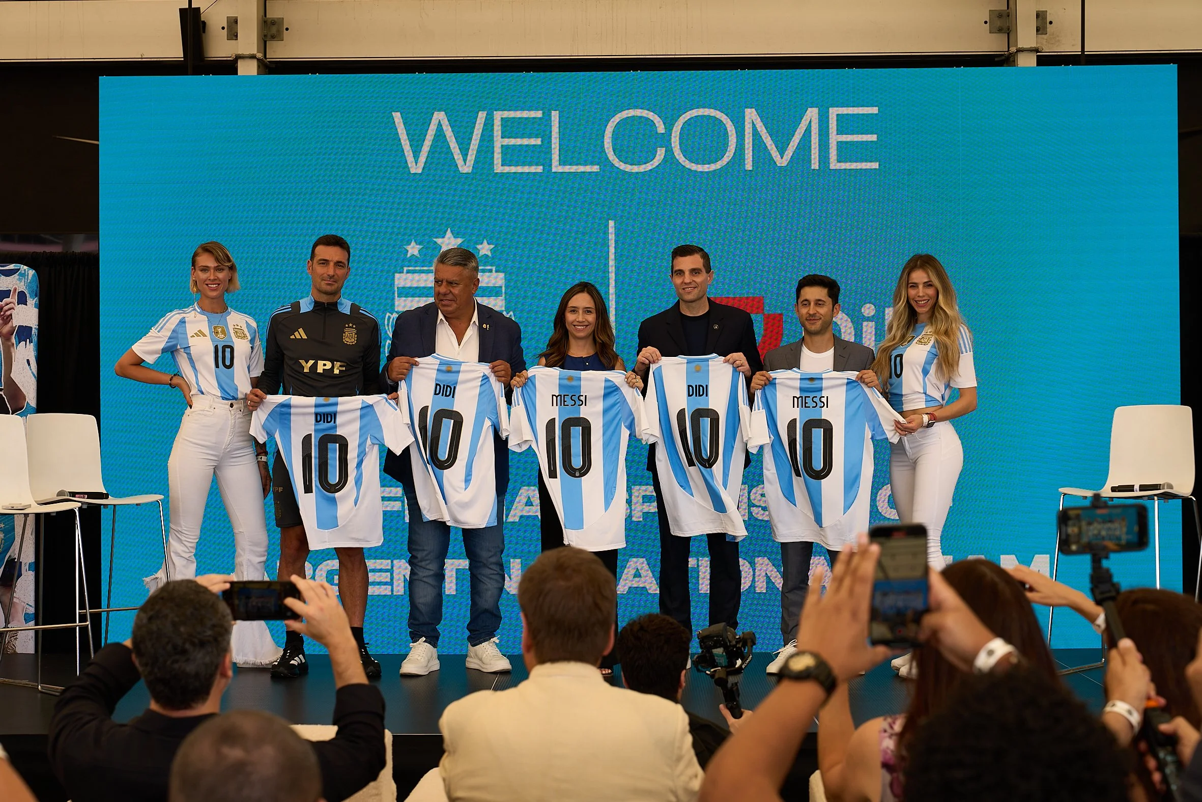 Group of people on stage holding Argentina soccer jerseys with the number 10, during a welcoming event, with an audience taking photos in front of a large blue screen display that reads 'WELCOME'.