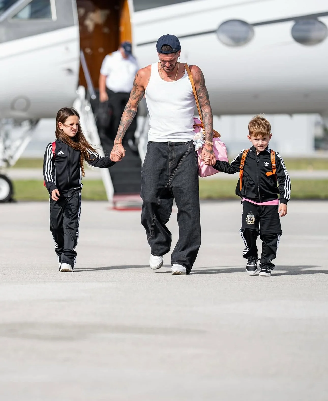 A man with tattoos, wearing a white tank top, dark baggy pants, and a navy cap, walking on the tarmac holding hands with two children, a girl and a boy, in black Adidas tracksuits, near an airplane.