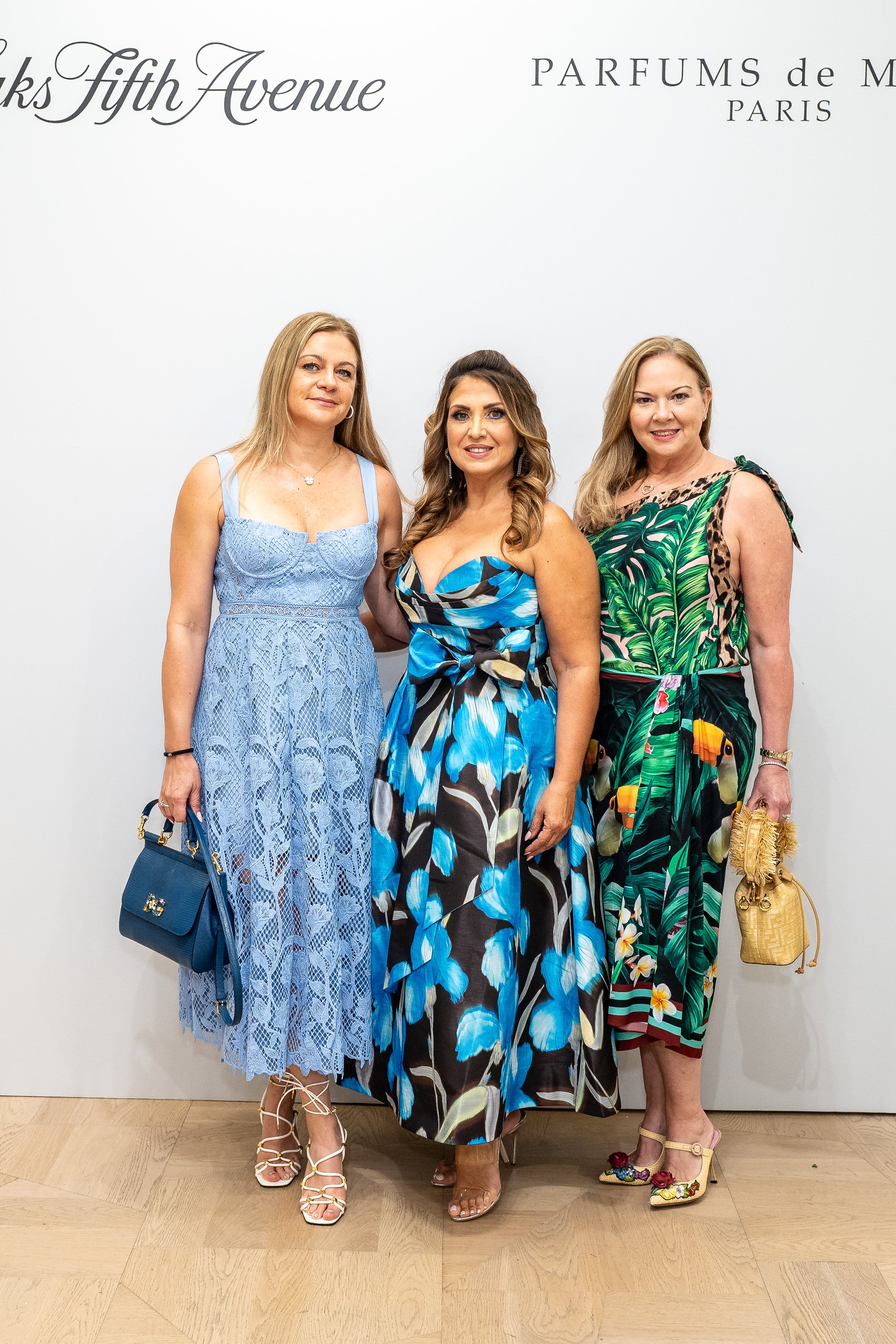 Three women standing together in front of a white wall with writing, dressed in colorful dresses and holding handbags, smiling at the camera.